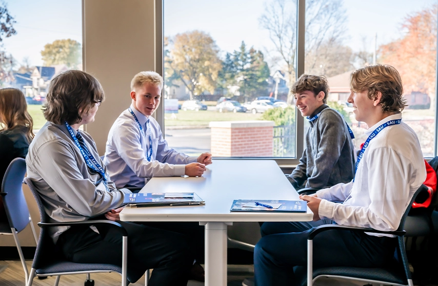 Four students talk at a table next to a window inside.