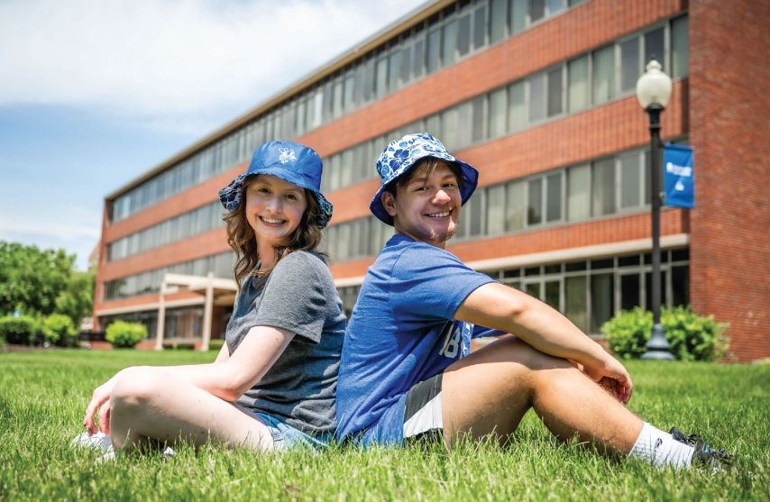 Two students wearing bucket hats smiling and sitting back to back.