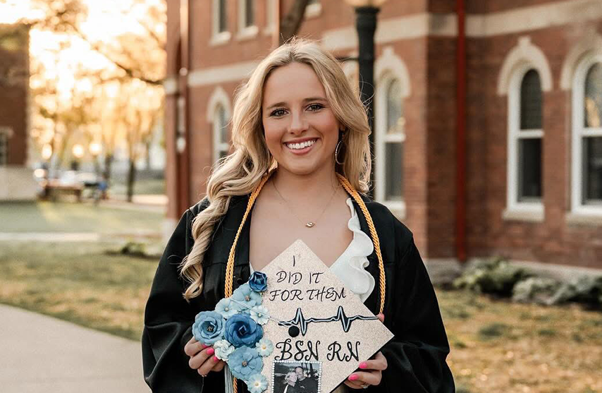 Abby Messina holds up a decorated graduation cap and smiles at the camera.