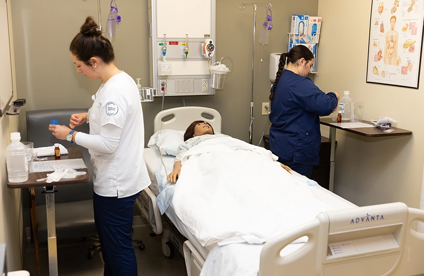 Two nursing students work together in a hospital room over a practice mannequin.