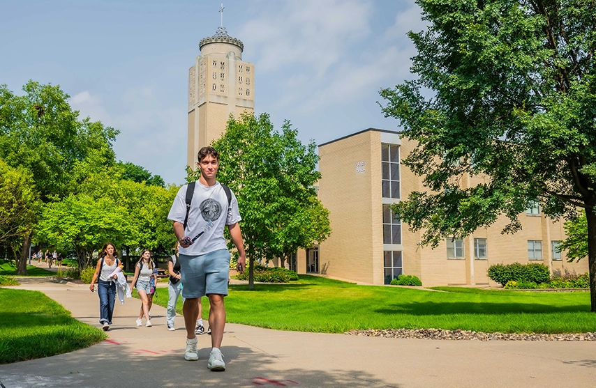 A group of St. Ambrose students walk outdoors past Christ the King Chapel on a sunny day.
