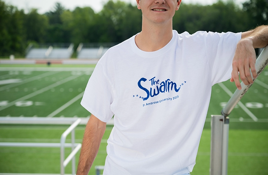 A student stands on a sports field while wearing a white shirt with blue text reading 