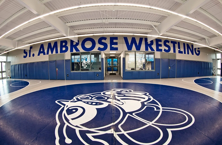 The blue and white wrestling gym at SAU with the SAU Bee on the floor and blue St. Ambrose Wrestling on  the rafters.