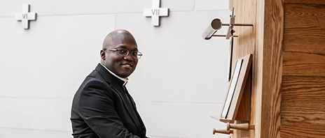 Fr. Eric Koami '25 smiles into the camera as he sits at the piano in Christ the King Chapel at St. Ambrose University.
