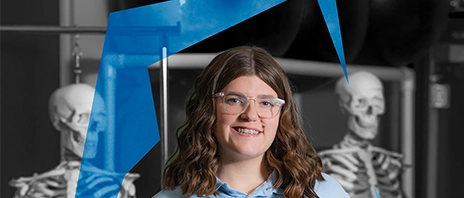 Sara Haller stands among educational skeletons, she smiles into the camera as the Ambrosian Arc surrounds her.