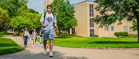 A group of St. Ambrose students walk outdoors past Christ the King Chapel on a sunny day.