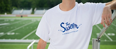 A student stands on a sports field while wearing a white shirt with blue text reading 