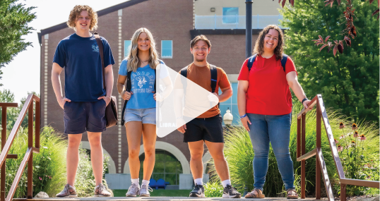 Four students standing outdoors in front of the SAU Library, smiling.