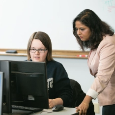 A woman helping a student at a computer in a classroom.