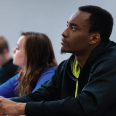 A diverse group of students sitting at desks in a classroom, listening to the teacher and taking notes.