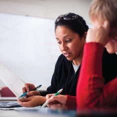 Two women in a classroom reviewing documents while a man is at a whiteboard in the background.