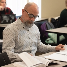 A man with glasses and a beard sitting at a desk with books and papers.