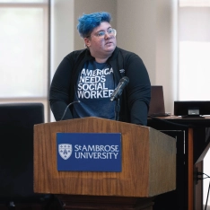 A person with blue hair standing at a podium, addressing an audience.