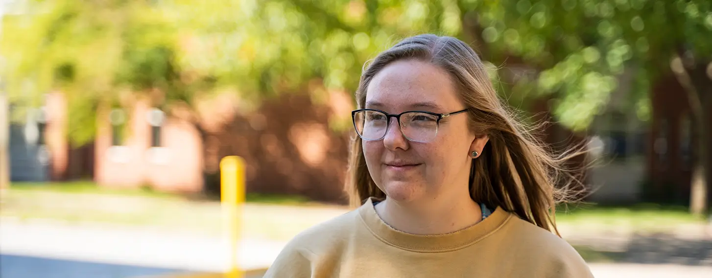 A girl with glasses and a gold sweatshirt walking outside on a sunny day.