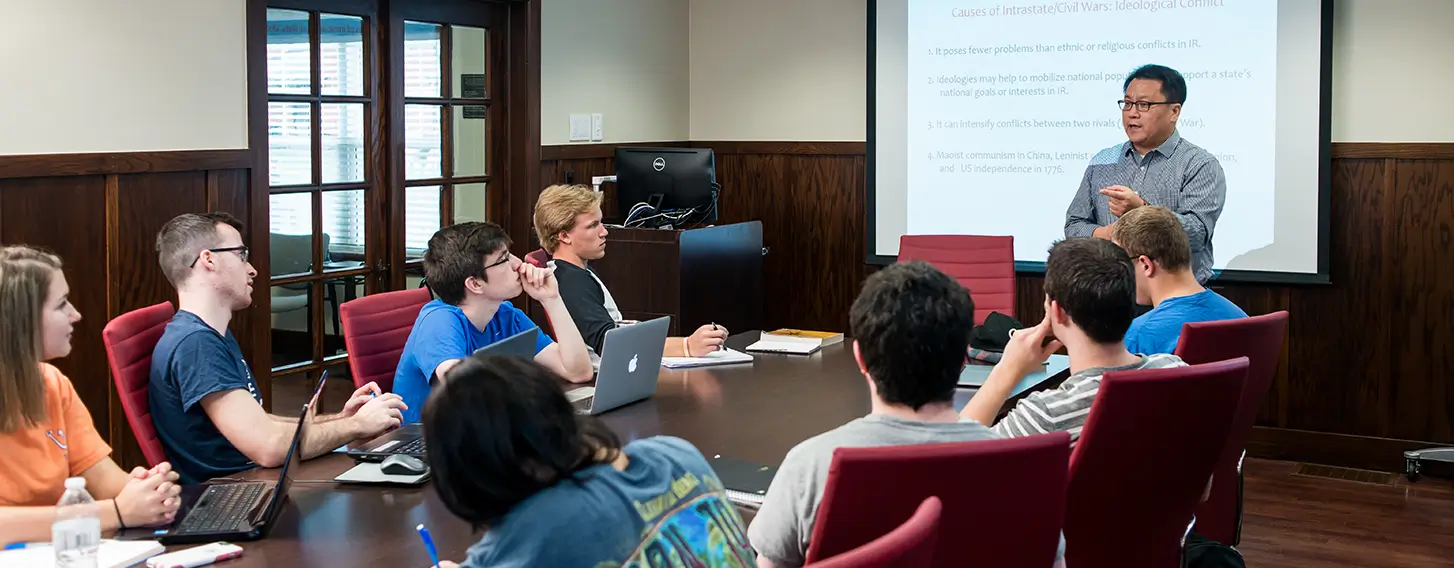 A group of students sitting around a boardroom style room during a presentation.