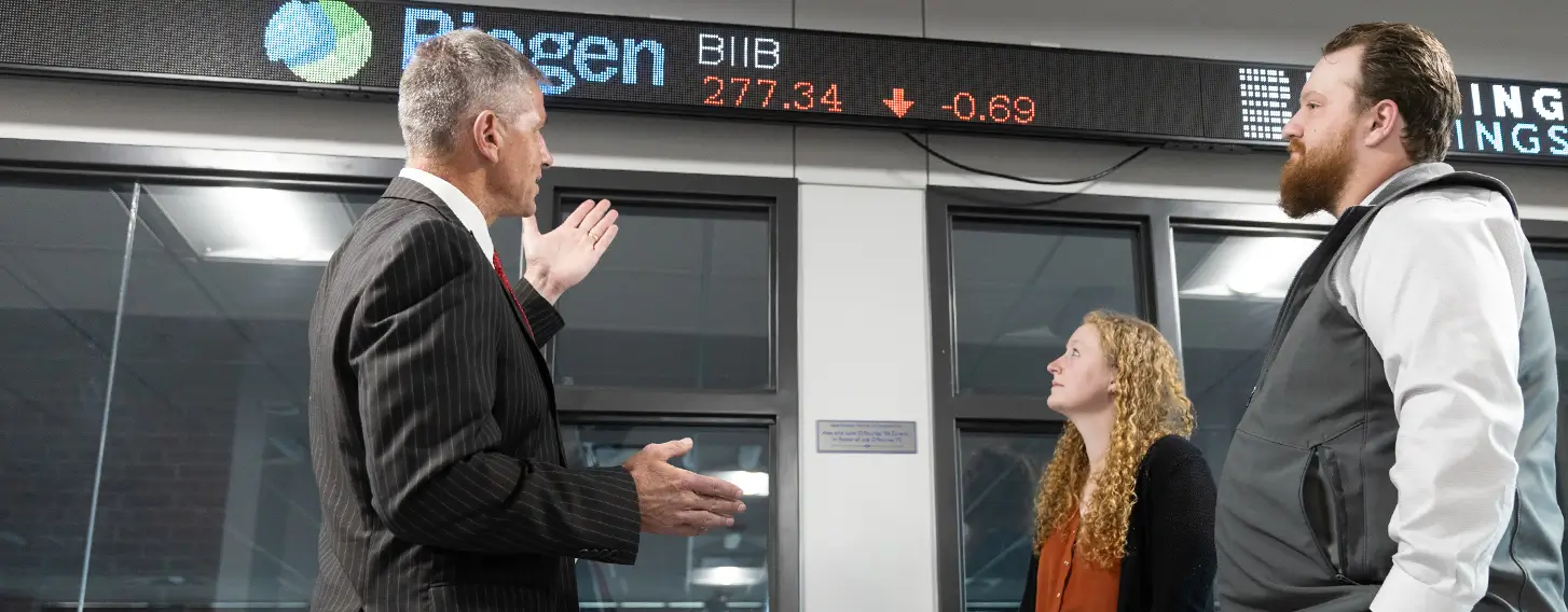 Three people standing in front of a glass wall with a stock market ticker displayed acroos the top of the wall.