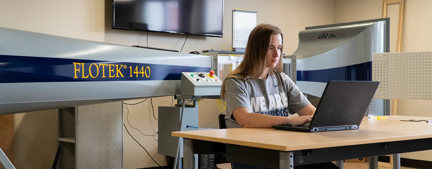 A female engineer sitting at a computer with a large wind tunnel in the background.