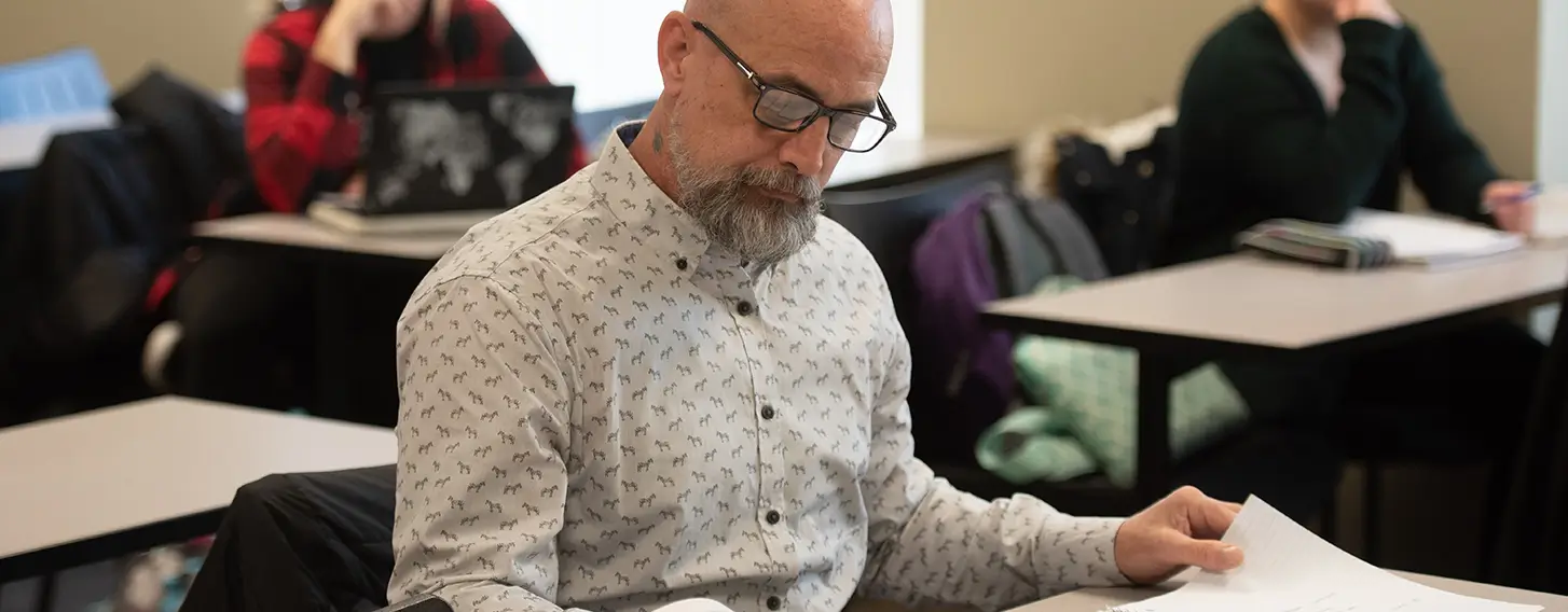 A man with glasses and a beard sitting at a desk with books and papers.