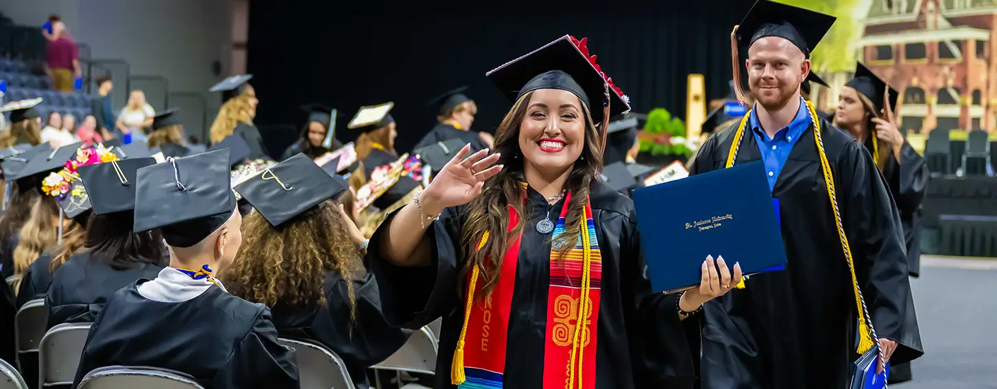 A woman in a graduation gown and cap smiling and waving during a graduation ceremony.