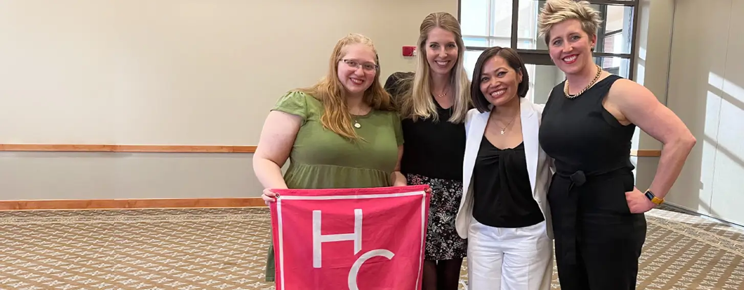 Four students posing with a pink HerCampus club flag.