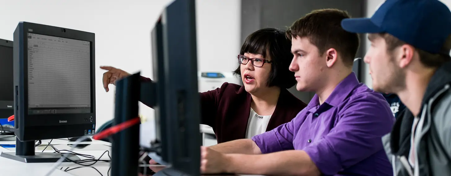 Students focused on computers in a classroom with their professor pointing something out on the screen.