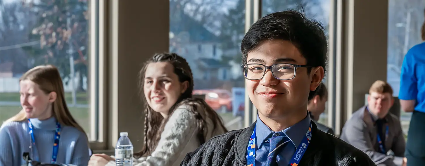 A young man sitting at a table with other students in the background in a classroom setting.