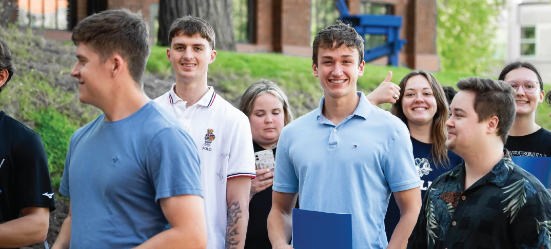 Students smiling at the camera and waiting in a line outdoors on a sunny day.