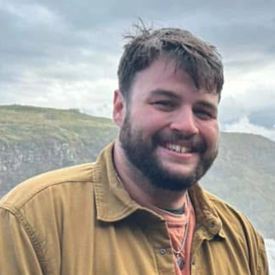 Luke Matthews smiles into the camera against a scenic cliffside.