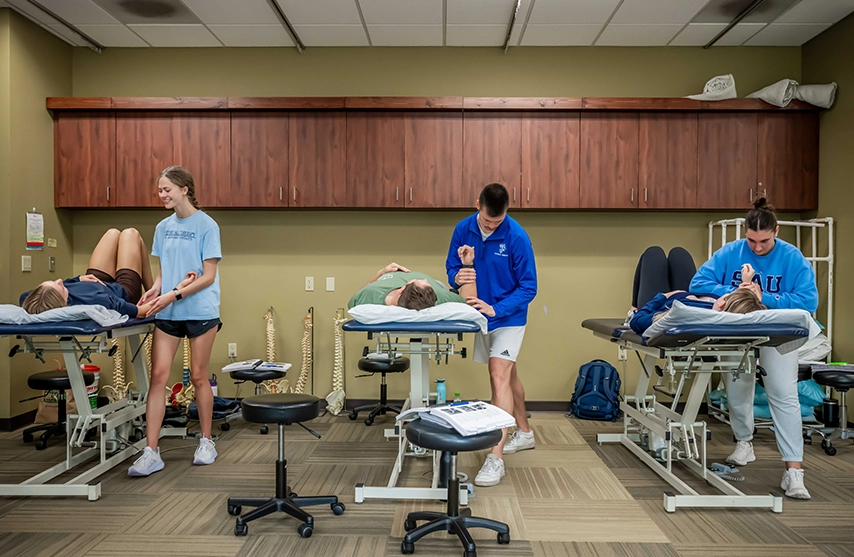 Physical therapy students practice manual therapy techniques with patients on treatment tables in a clinical lab.