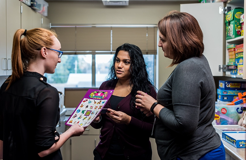 Three women talk while holding papers.