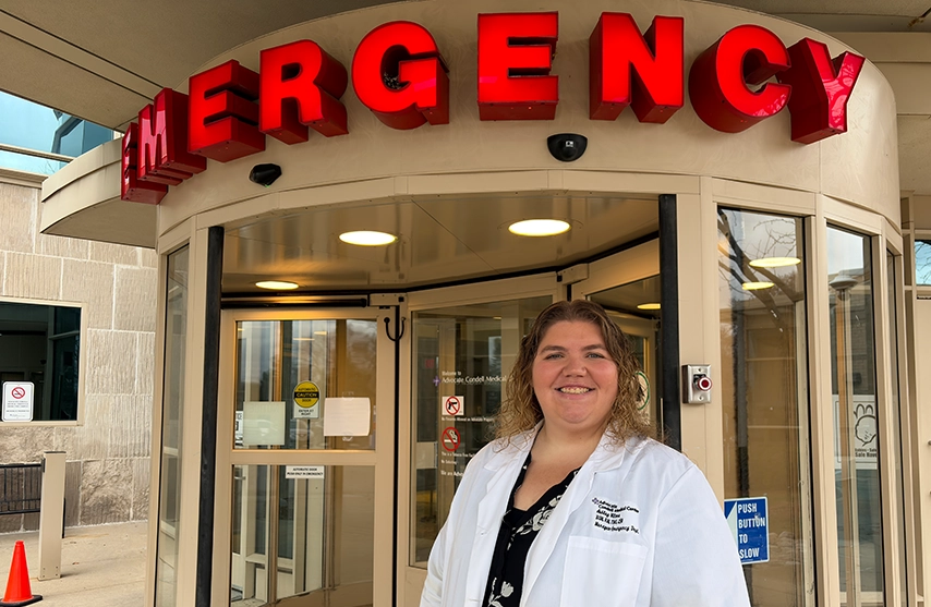 Ashley Vilona, '14, smiles in her doctor's coat while standing in front of Emergency Room revolving doors.