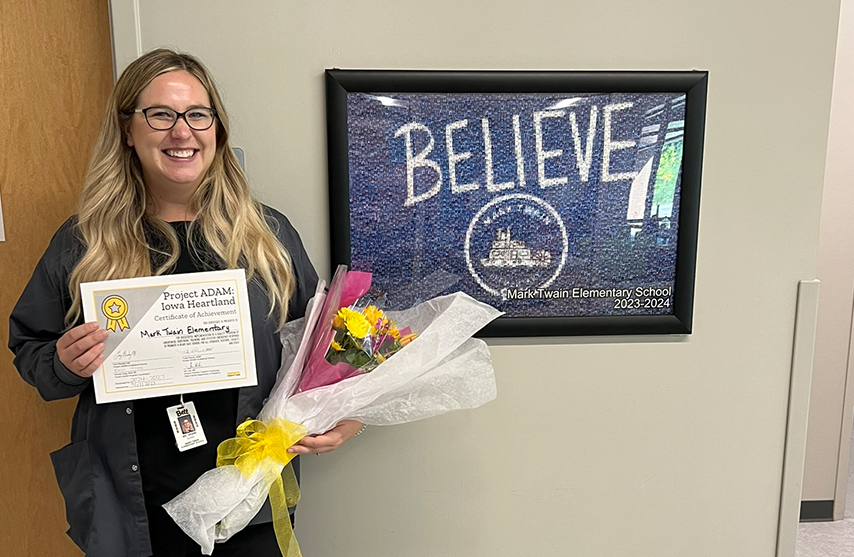 Jordan Drane stands in front of a framed blue photo with the word believe. She holds follows and a certificate.