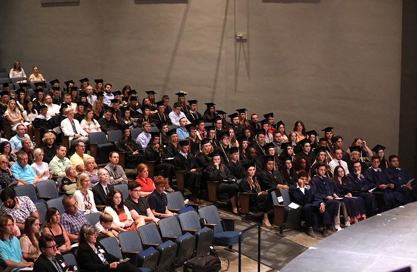 An audience of students in graduation regalia and parents face a stage out of frame.