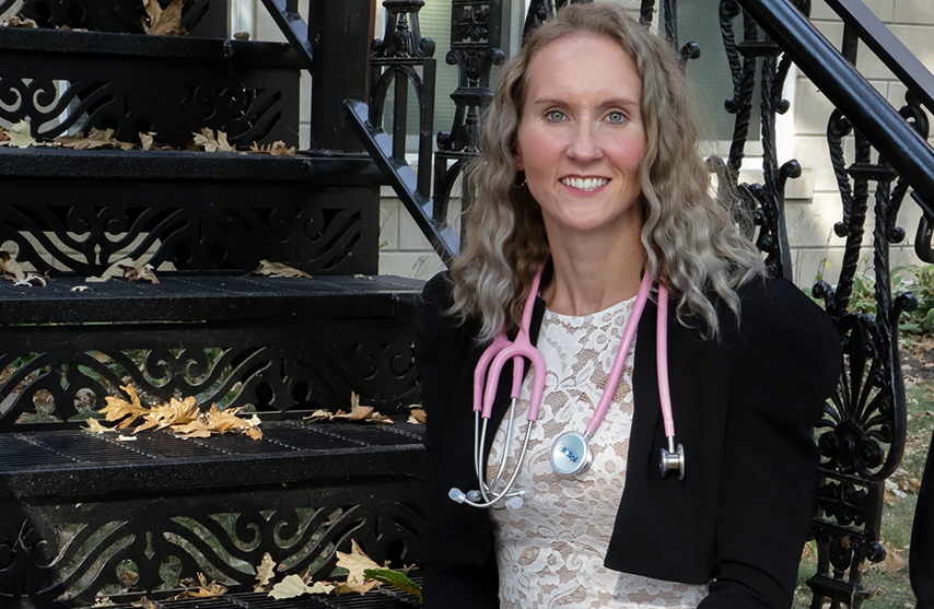 Sarah Burr sits on metal stairs with a pink stethoscope and smiles at the camera.