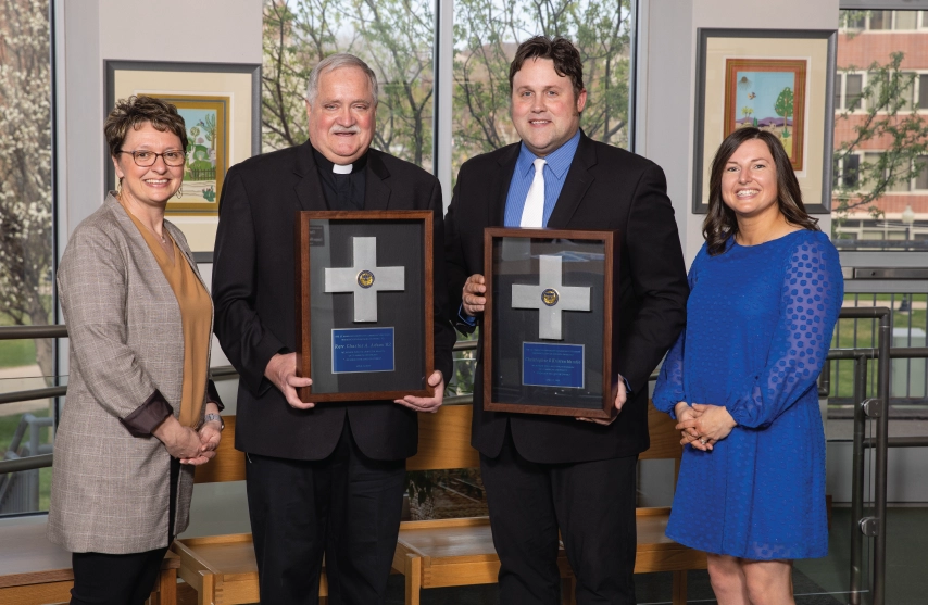 Four people posing together. The two men in the middle are each holding an award.