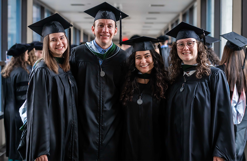 A group of four graduates smile at the camera while dressed in commencement regalia.