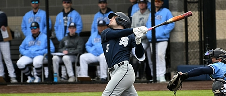 St. Ambrose baseball player follows through on a swing at home plate as teammates watch from the dugout in the background.
