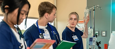Students of St. Ambrose University's nursing program gather around a hospital bed with folders and notebooks.