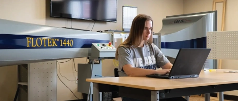 A female engineer sitting at a computer with a large wind tunnel in the background.