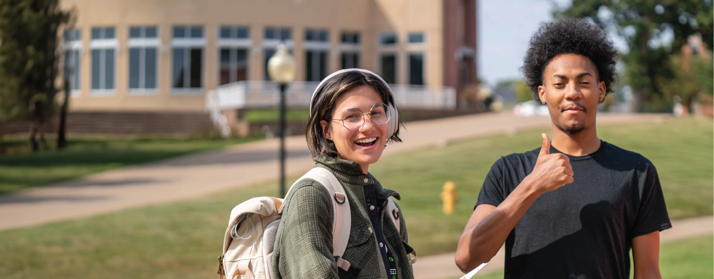 Two students standing outdoors, smiling, with one giving a thumbs up sign.