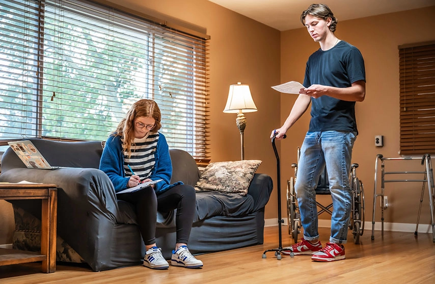 A young woman sits on a couch while writing on a clipboard, a young man stands while reading from a paper and holding a cane.