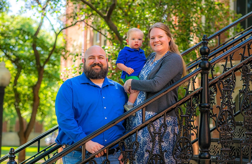 A man in a blue shirt, a woman in a blue dress holding a baby in a blue shirt stand on stairs and smile at the camera.