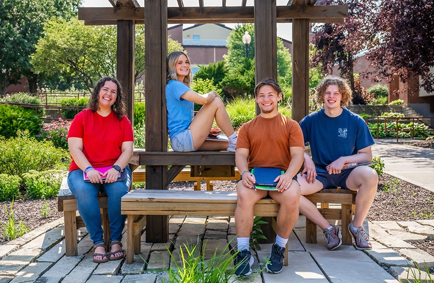 Four students sitting outside at a wooden patio. They smile at the camera.
