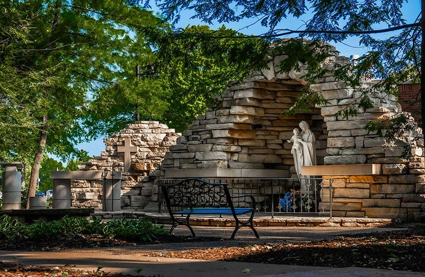 A bench under a tree faces a stone wall and statue.